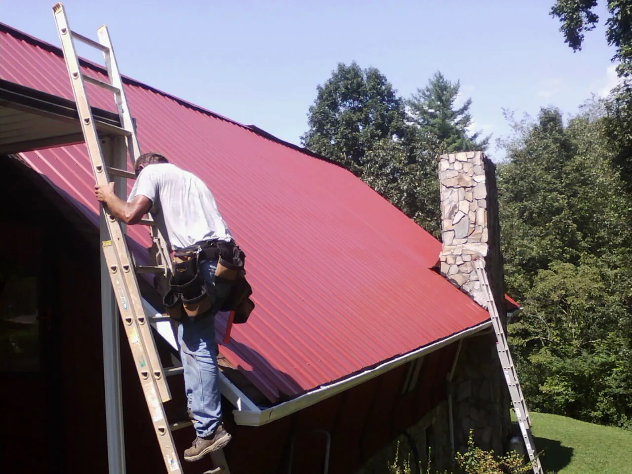 Roofer on a ladder beside a red metal roof and stone chimney — Soffit & Fascia Repair by Roofing Brothers in Mona